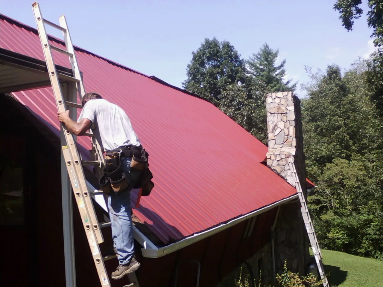 Roofer on a ladder beside a red metal roof and stone chimney — Soffit & Fascia Repair by Roofing Brothers in Fruita